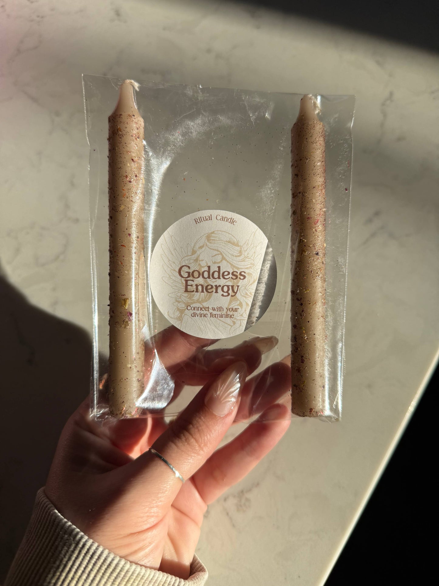 Two candles in clear packaging with a label reading 'Goddess Energy' held by a hand.